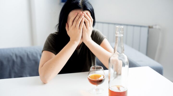 Woman sitting at a table with her head in her hands, a glass and bottle of liquor in front of her.