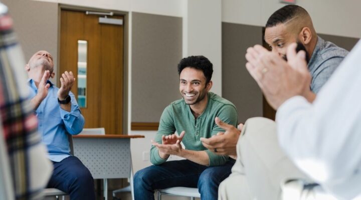 Group of men sitting in a circle during a meeting, smiling and clapping in celebration, with one man at the center looking joyful.