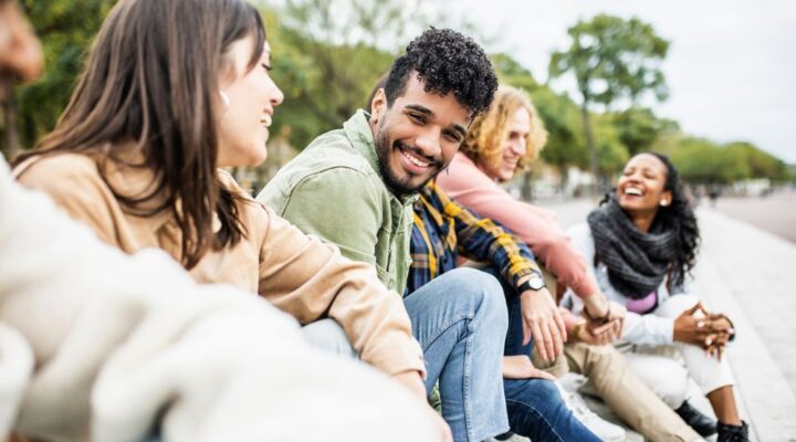 Group of diverse young adults sitting outdoors, smiling and talking together in a relaxed, friendly atmosphere.