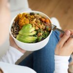 Person holding a bowl of healthy food, including avocado, greens, beans, and roasted vegetables, representing balanced nutrition and wellness.