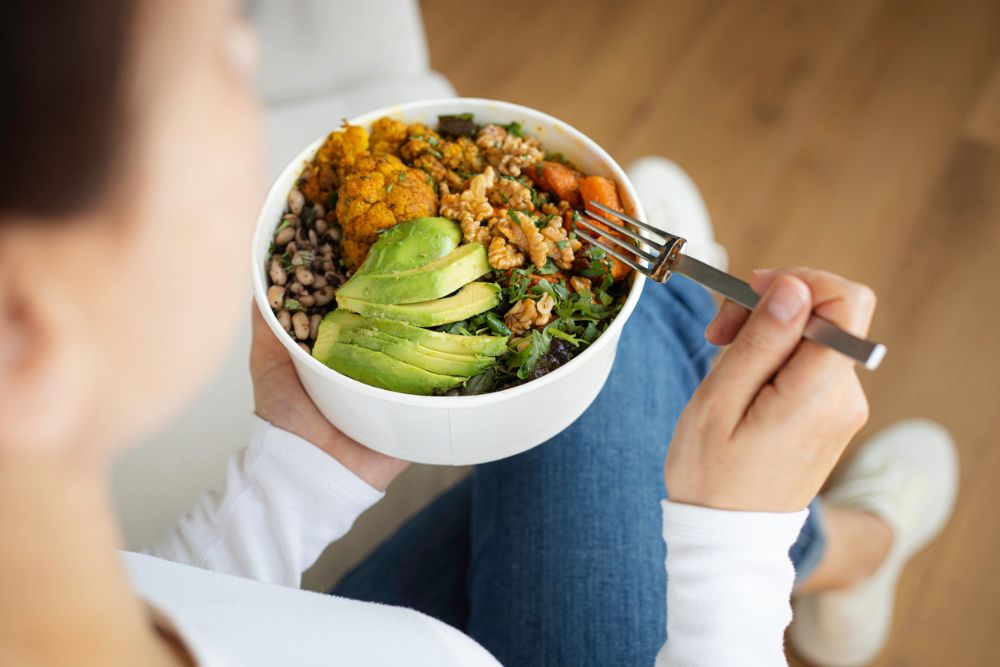Person holding a bowl of healthy food, including avocado, greens, beans, and roasted vegetables, representing balanced nutrition and wellness.