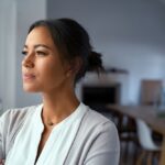 A woman stands indoors, gazing thoughtfully out a window with a calm, serious expression, suggesting reflection, solitude, or emotional resilience.