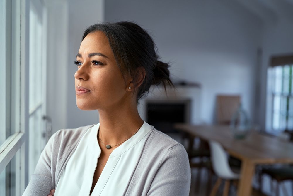 A woman stands indoors, gazing thoughtfully out a window with a calm, serious expression, suggesting reflection, solitude, or emotional resilience.