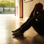 A man sits on the floor in a dimly lit hallway, leaning against the wall with his elbows on his knees and his hands holding his head. He appears distressed and overwhelmed.