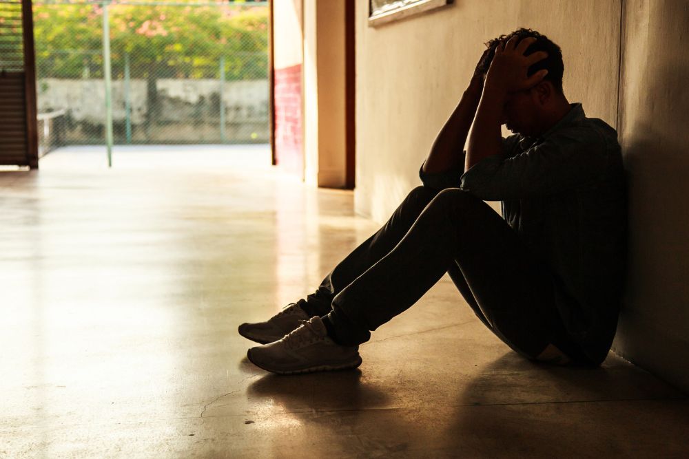 A man sits on the floor in a dimly lit hallway, leaning against the wall with his elbows on his knees and his hands holding his head. He appears distressed and overwhelmed.