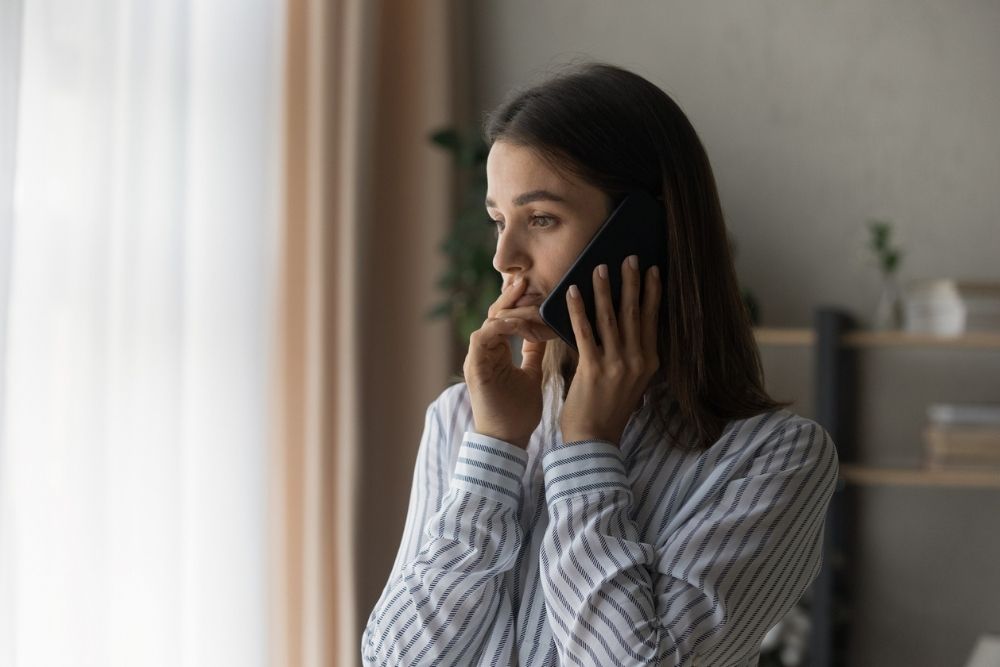 Woman standing indoors, holding a phone to her ear with a thoughtful, concerned expression, wearing a striped blouse.