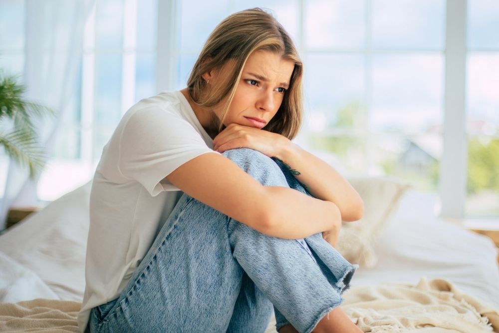 A young woman sits on her bed, hugging her knees, looking sad and thoughtful. The bright room contrasts with her emotional expression, conveying anxiety or loneliness. Mental health and depression.