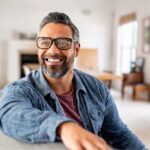 Confident middle-aged man with glasses and a salt-and-pepper beard smiling while sitting in a bright, modern living room, radiating positivity and self-assurance.