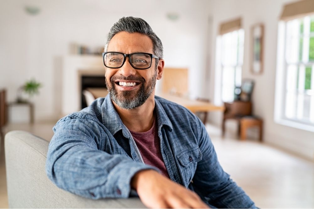 Confident middle-aged man with glasses and a salt-and-pepper beard smiling while sitting in a bright, modern living room, radiating positivity and self-assurance.