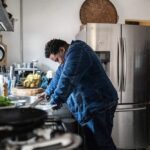 Person leaning over a kitchen counter with their head down, appearing tired or overwhelmed, in a home setting.