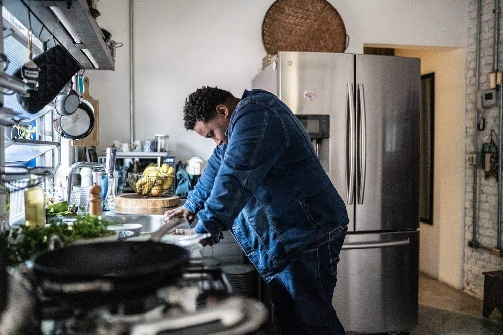 Person leaning over a kitchen counter with their head down, appearing tired or overwhelmed, in a home setting.