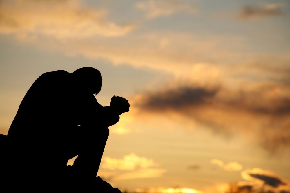 A person sits outdoors at sunset with their head bowed and hands clasped, appearing deep in thought or prayer, symbolizing struggle or reflection.