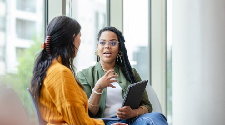Two women sit and talk near large windows; one holds a tablet and gestures while speaking, suggesting a supportive conversation.