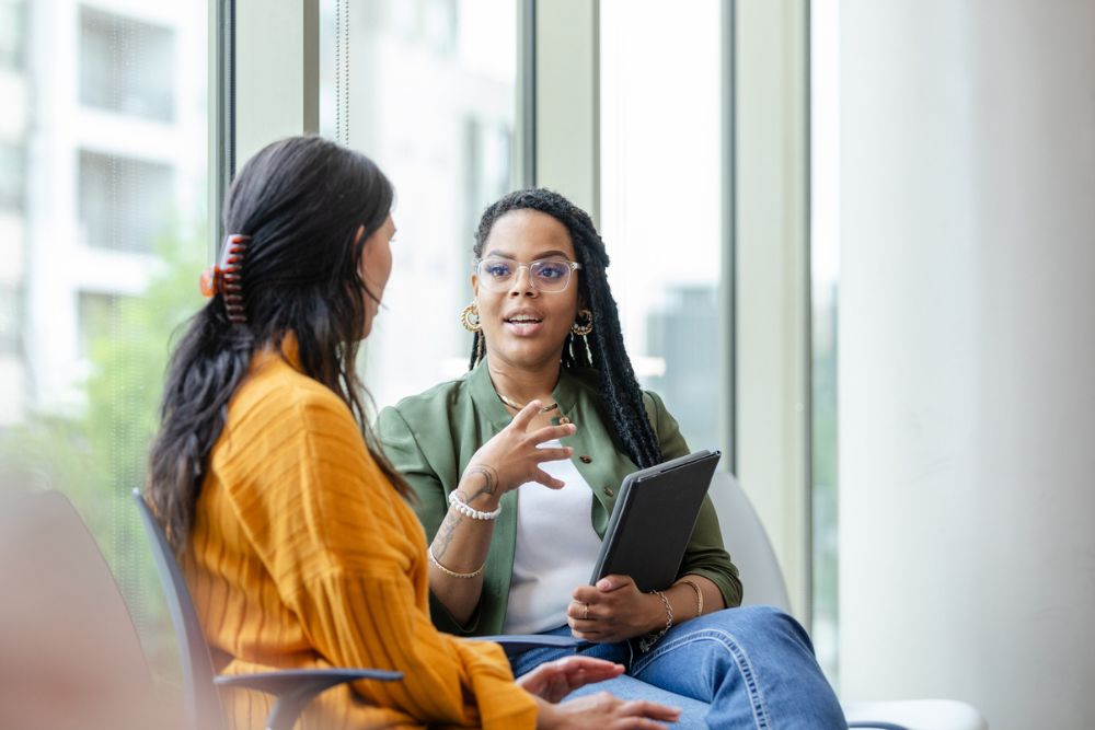 Two women sit and talk near large windows; one holds a tablet and gestures while speaking, suggesting a supportive conversation.
