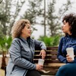 Two women sitting on a park bench holding coffee cups, engaged in a thoughtful conversation on a cloudy day surrounded by trees.