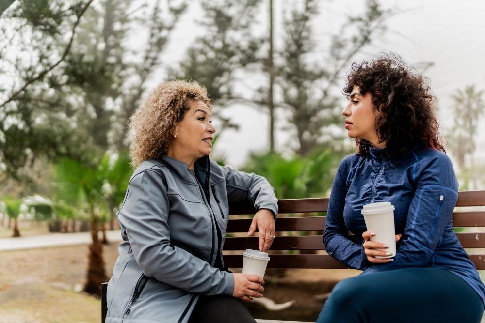 Two women sitting on a park bench holding coffee cups, engaged in a thoughtful conversation on a cloudy day surrounded by trees.