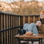 Young woman in knitted sweater and hat drinking tea and eating fresh croissants on cozy balcony of a wooden country house on autumn day.
