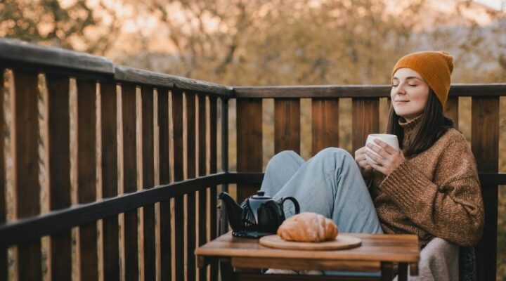 Young woman in knitted sweater and hat drinking tea and eating fresh croissants on cozy balcony of a wooden country house on autumn day.