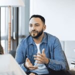 A man sitting in a therapy session gestures while speaking, facing a therapist in a bright, modern office setting.