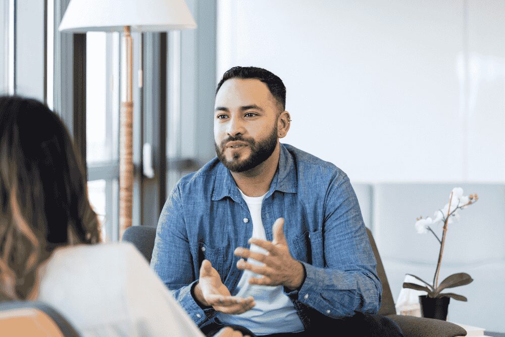 A man sitting in a therapy session gestures while speaking, facing a therapist in a bright, modern office setting.