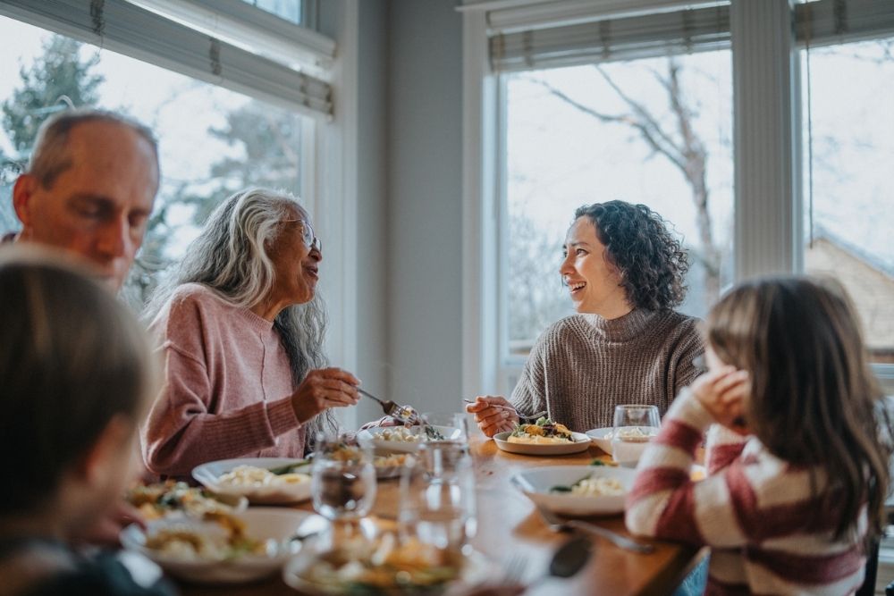 A family gathered around a dining table, sharing a meal and laughing together near large windows on a winter day.