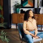 A woman sits cross-legged on a chair with her eyes closed, practicing mindfulness in a calm, modern living space filled with indoor plants and warm natural light.