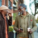 Two men stand outdoors laughing together, holding coffee cups and chatting, reflecting a warm, supportive friendship in a neighborhood setting.