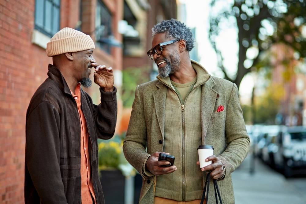 Two men stand outdoors laughing together, holding coffee cups and chatting, reflecting a warm, supportive friendship in a neighborhood setting.