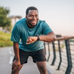 Man in athletic wear smiling and checking his smartwatch on a paved path - working to build a healthier lifestyle for himself.