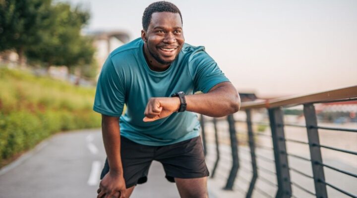Man in athletic wear smiling and checking his smartwatch on a paved path - working to build a healthier lifestyle for himself.