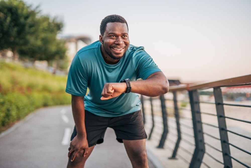 Man in athletic wear smiling and checking his smartwatch on a paved path - working to build a healthier lifestyle for himself.
