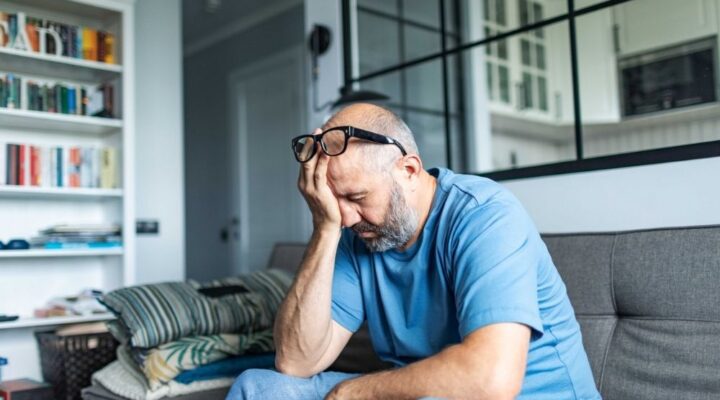 Person sitting on a couch with head in hand, appearing stressed or overwhelmed in a quiet home setting.