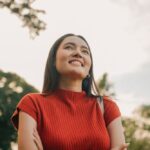 Smiling woman in a red shirt stands outdoors with her arms crossed, looking up confidently against a bright sky.