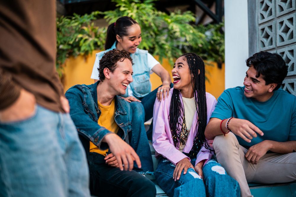 Stone River - The Language You Use Matters - Edited. Friends sitting on a bench in an outdoor environment smile and laugh with one another in conversation.